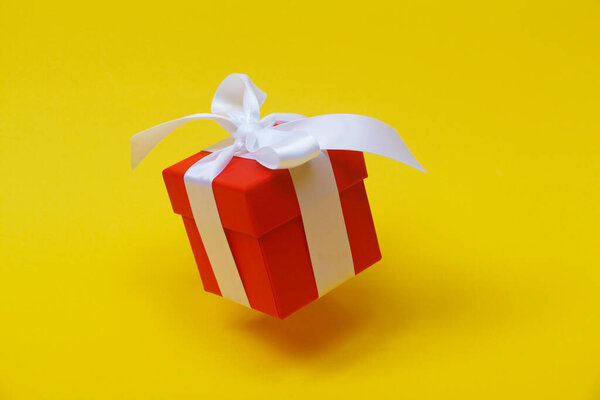 Красная подарочная коробка с белым бантом на желтом фоне. Коробка закрыта крышкой. Подарок в воздухе
.
