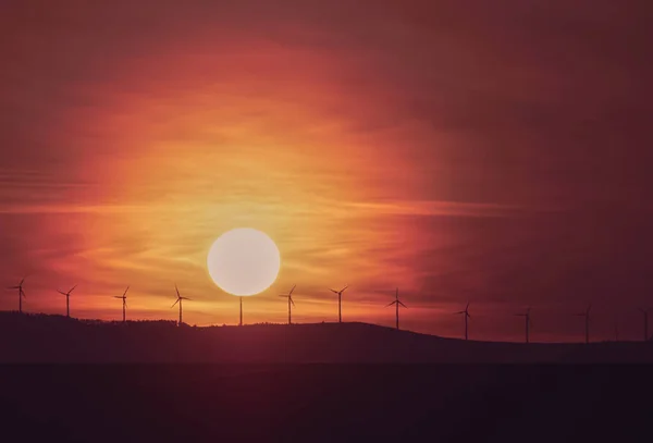 Sunrise with wind turbines on the hill and sun and clouds in the background.