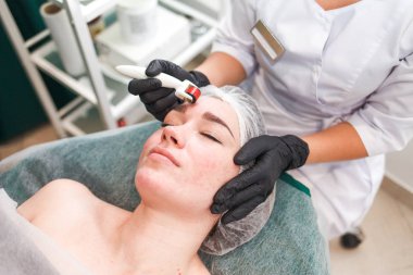 Doctor cosmetologist makes facial massage procedure using a dermo roller. Woman in beauty salon during mesotherapy procedure with mesoscooter clipart
