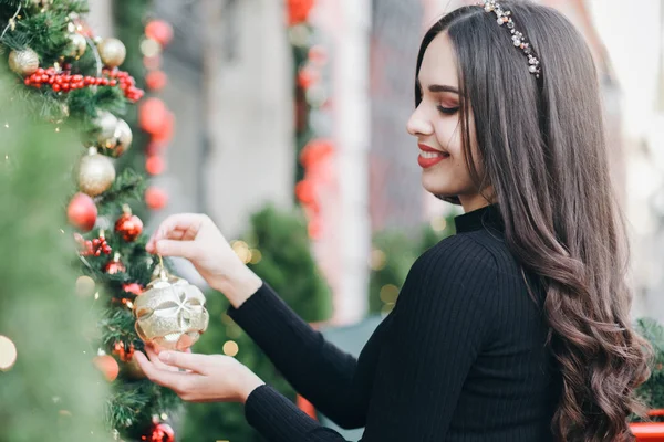 Beautiful young girl hangs balls on a Christmas tree near the store. Cute smiling girl decorates a Christmas tree