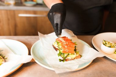 Ellerinizi pişirin ve sandviç yapın. Sağlıklı vejetaryen bruschetaları siyah eldivenlerle hazırlamak. Yumuşak peynirli sandviç, roka, salatalık ve susamlı somon.