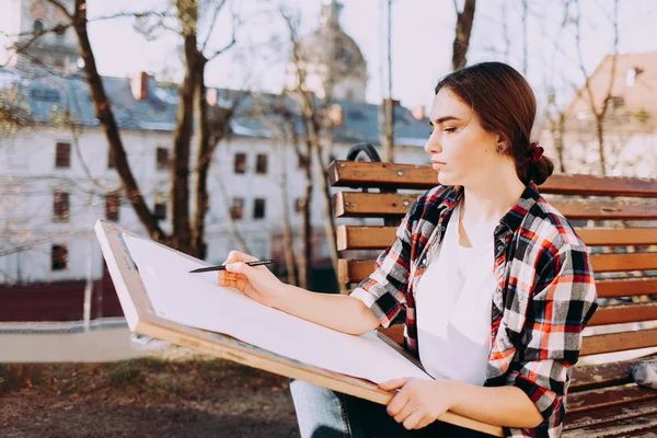Young woman artist draws a picture while sitting on a bench. Painter artist holds a wooden tablet for painting and a brush in his hand