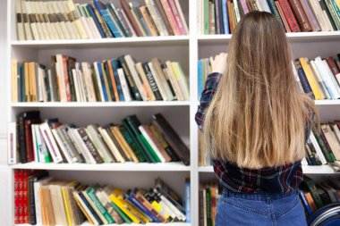 Gözlüklü sevimli öğrenci kütüphane kitaplığından bir kitap alıyor. Genç bayan kitapların arasında doğru edebiyatı arıyor.