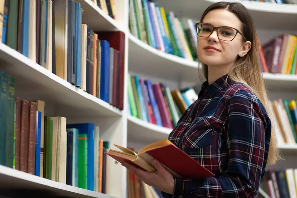 手里拿着一本书的青少年站在图书馆里 一个戴眼镜的年轻学生在看书的画像 — 图库照片