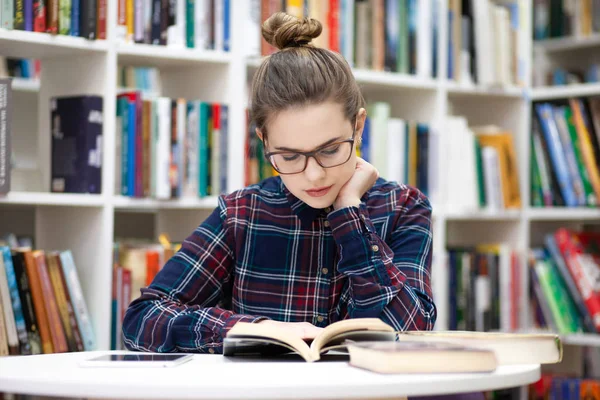 Girl sitting in the library. Young student in glasses in a checkered shirt sits in a public library and reading a book. Woman likes to develop reading books