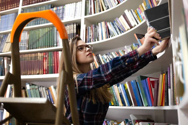 Cute student in glasses takes a book from the library bookshelf. Young lady is looking for the right literature among many books