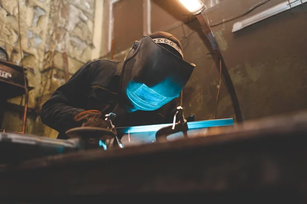 Worker in a welder mask works in a workshop for welding iron. Man makes iron products. Guy works with a welding machine using a mask to protect his eyes from dangerous rays