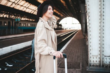 Tren istasyonunda gülümseyen güzel bir kadın. Trenini bekleyen bir bavulla tren istasyonunda duran şapkalı ve paltolu pozitif turist kadın..