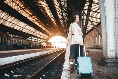 Tren istasyonunda gülümseyen güzel bir kadın. Trenini bekleyen bir bavulla tren istasyonunda duran şapkalı ve paltolu pozitif turist kadın.