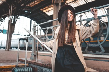 Portrait of a positive beautiful stylish passenger woman in coat and hat at the railway station