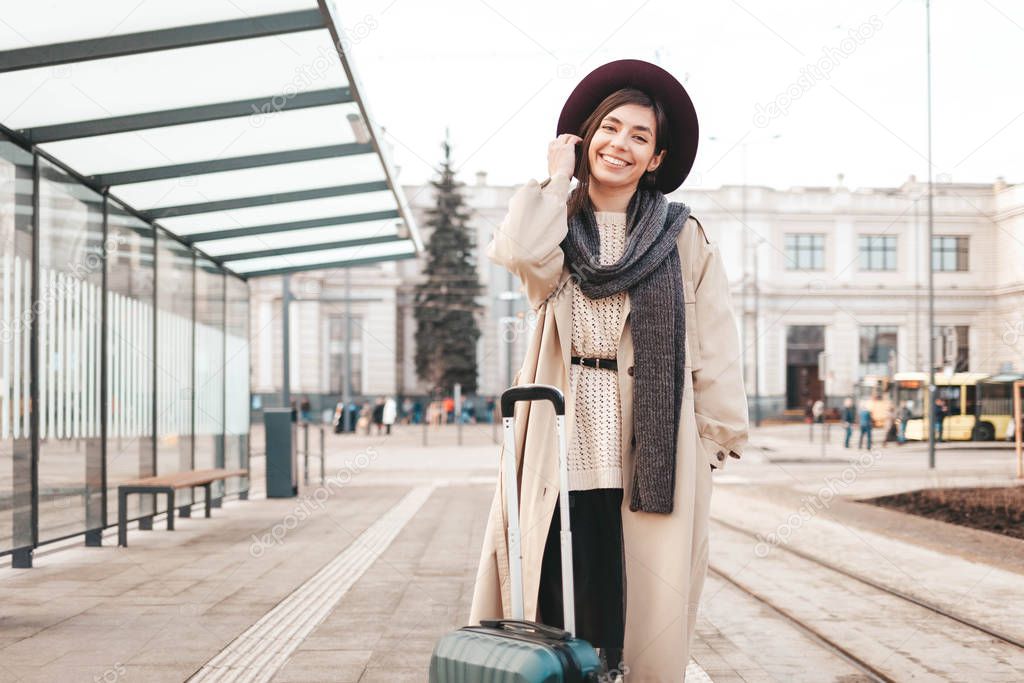 Stylish girl with a suitcase stands at a city stop awaiting a tram