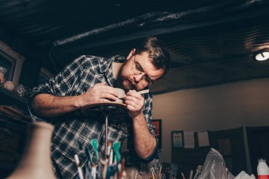 Sakallı adam çömlek atölyesinde çalışıyor, kilden bir bardak yapma sürecine odaklanıyor.