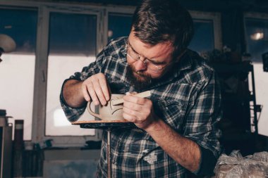 Sakallı adam çömlek atölyesinde çalışıyor, kilden bir bardak yapma sürecine odaklanıyor.