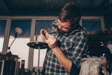 Sakallı adam çömlek atölyesinde çalışıyor, kilden bir bardak yapma sürecine odaklanıyor.