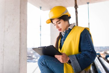 Koruyucu başlık takan genç bir mühendis kız inşaat alanında kağıt dolduruyor.