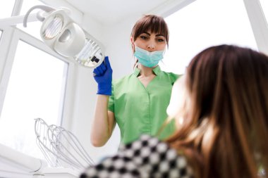 Kız dişçi randevusunda sandalyede yatıyor. Dişçi doktor hastaların dişlerine lamba takıyor.