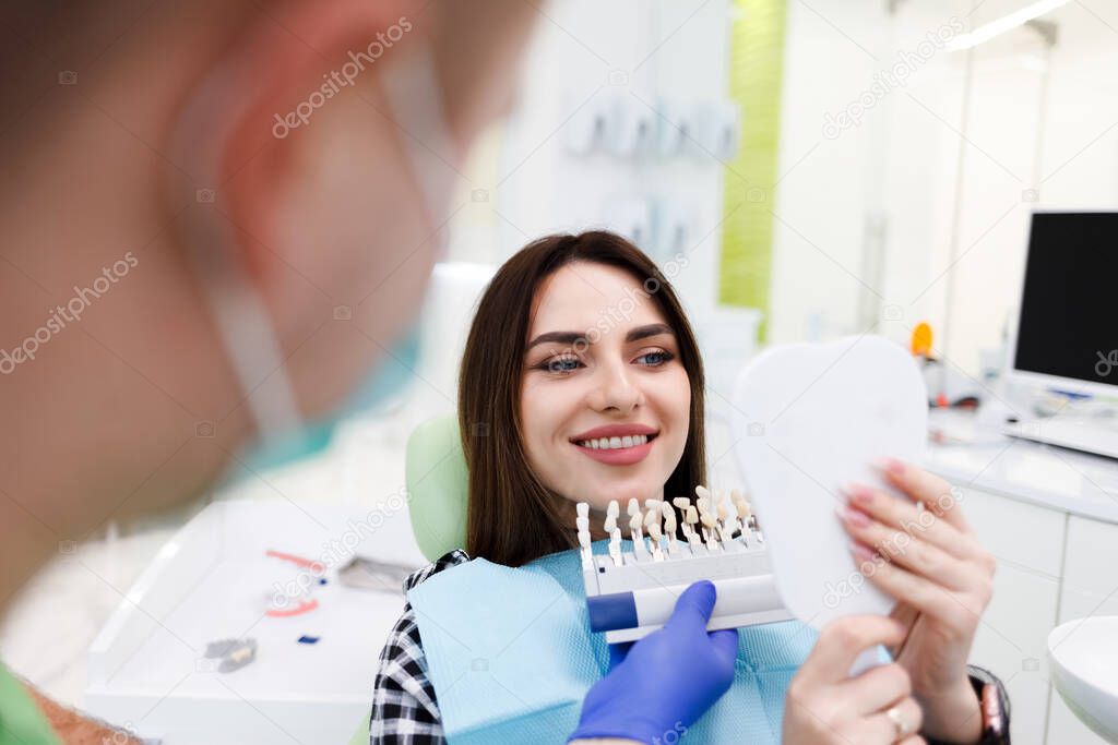 Patient in a dental office holds a palette with shades of teeth and looks in the mirror