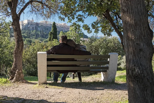 Genç çift parkta bir bankta oturuyor. Atina Akropolünün panoramik görüntüsü