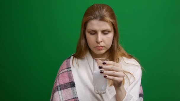 Frau trinkt eine Brausetablette. Die Pille fällt und löst sich mit Blasen auf. Rote Haare. COVID Brausetablette in einem Glas Wasser auf grünem Hintergrund. — Stockvideo