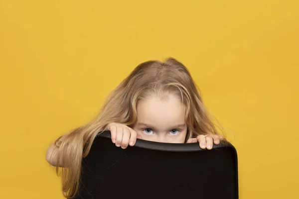 Funny and pretty child girl with long fair hair peeks out from behind a chair. — Stock Photo, Image