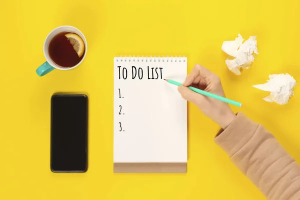 To do list. Mint Tea cup and notebook with to do list on bright yellow desk from above, planning and design concept.