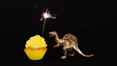 Siyah arka planda izole edilmiş parlak sarı bir kek üzerinde altın dinozor ve maytap. Mutlu yıllar konsepti. HD video. Doğum günü pastası ve oyuncak dinozor