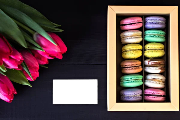 Box with macaroons and a bouquet of tulips and a card for your text on a dark background. — Stock Photo, Image
