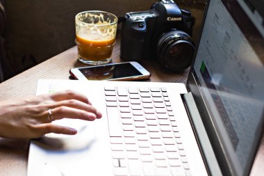 Kız bilgisayarda çalışıyor. Eller bilgisayar, fotoğraf makinesi ve kahve arka planda çalışıyor. Fotoğrafçı bir kafede iş başında.