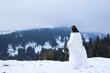 Kış düğün portresi. Kafasında çiçek ve pamuk çelenk olan güzel gelinin portresi, elinde buket, karlı havada kış ormanının arka planında beyaz bir elbise giyiyor.