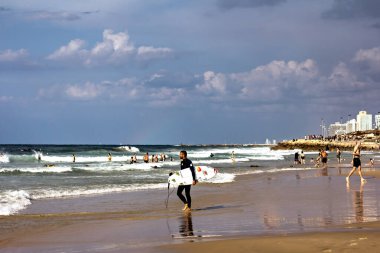 Tel Aviv, İsrail, Kasım 2019 sörfçü, sörf tahtası olan genç bir adam deniz kenarında yürüyor. Denizde sörfçü