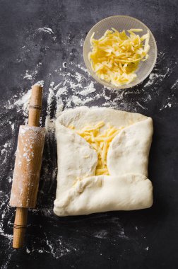 Siyah bir tahta üzerinde hamur, fırın un, rendelenmiş peynir ve haddeleme pin haddelenmiş.