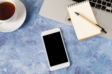 Ofis masasında siyah ekranlı, dizüstü bilgisayarlı, boş defterli, beyaz akıllı telefon ve bir fincan çay. Telefonun taklidi..