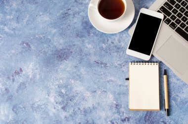Ofis masasında siyah ekranlı, dizüstü bilgisayarlı, boş defterli, beyaz akıllı telefon ve bir fincan çay. Telefonun taklidi..