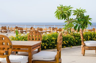 Tahta masalar ve terastaki rahat yazlık kafe denize bakan sandalyeler. Deniz kenarında güneşli bir yaz günü. Kafe ya da restoranın dışı.