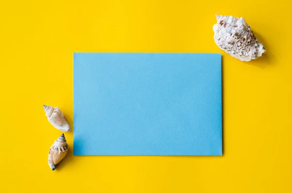 Pusty niebieski arkusz papieru na żółtym tle z muszlami. Tło letnie. — Zdjęcie stockowe