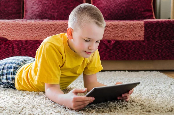 Distance learning online education. Schoolboy studying at home with digital tablet in hand and doing school homework. Boy looks at tablet, lying on floor.