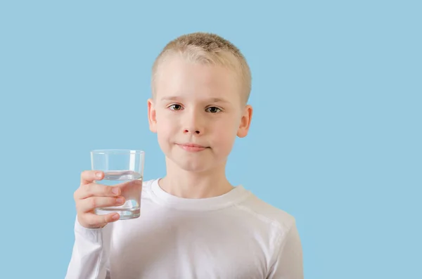 Dziecko trzyma szklankę wody w rękach. Blond mały chłopiec ze szklanką wody, patrząc w aparat. — Zdjęcie stockowe