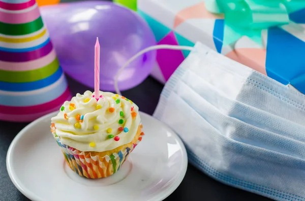 Quarantine birthday in isolation. Birthday cupcake with candle, face mask, gifts and holiday accessories. Social distance.