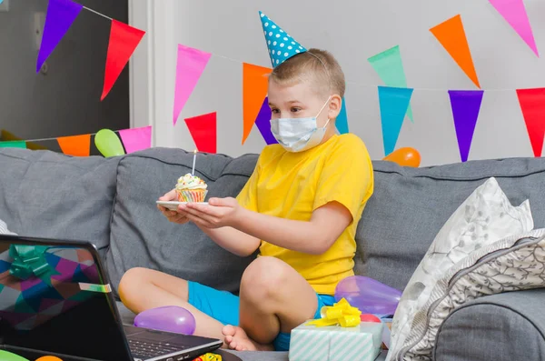 Happy boy in medicine face mask with holiday cupcake celebrates birthday by video call to laptop. Quarantine birthday online in isolation. Virtual event keeping social distance.