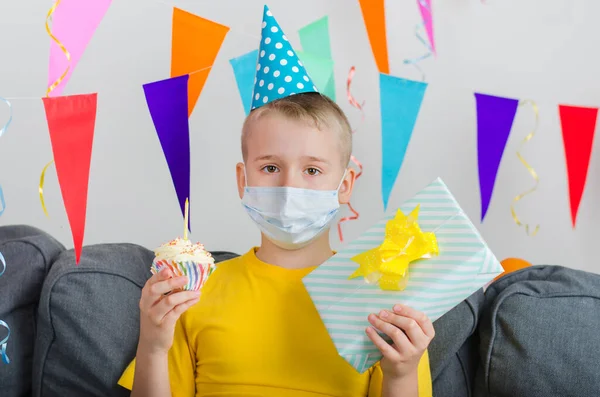 Sad boy in medicine face mask and festive cap with gifts in hand celebrates birthday. Quarantine birthday alone in isolation. Social distance.