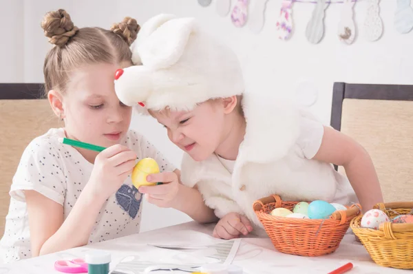 Påskhögtiden: Barn måla påskägg vid bordet. En pojke i en kanin kostym ser listigt på arbetet av storasyster. — Stockfoto