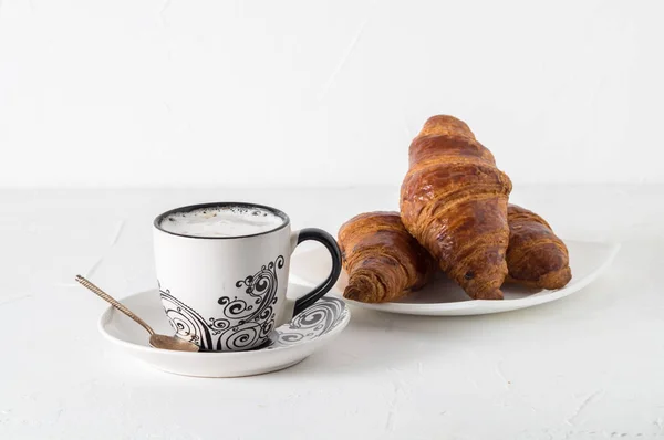 Coffee and pastries: a white coffee mug with a pattern, a silver spoon, fresh croissants on a saucer. — Stock Photo, Image