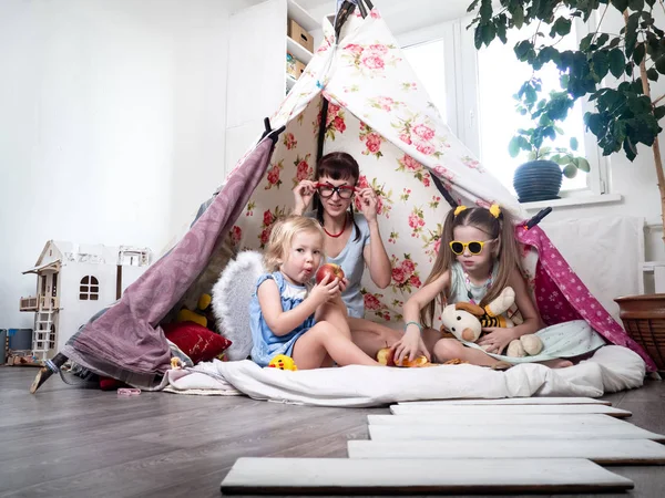 Tiempo en familia: Mamá y algunas de las hermanas niños juegan en casa en una tienda de campaña casera para niños . —  Fotos de Stock