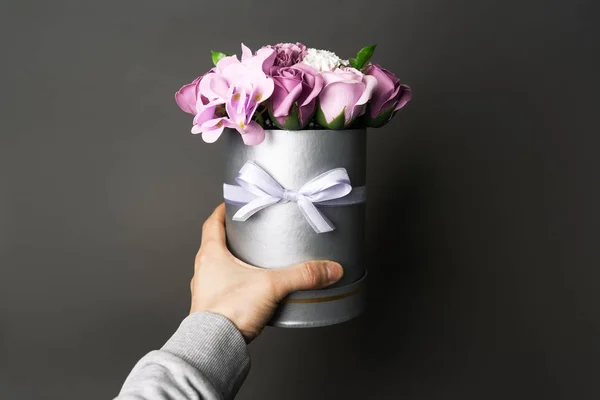 Flores en flor: Un ramo de rosas lila y blanca en una caja gris redonda en una mano masculina . — Foto de Stock