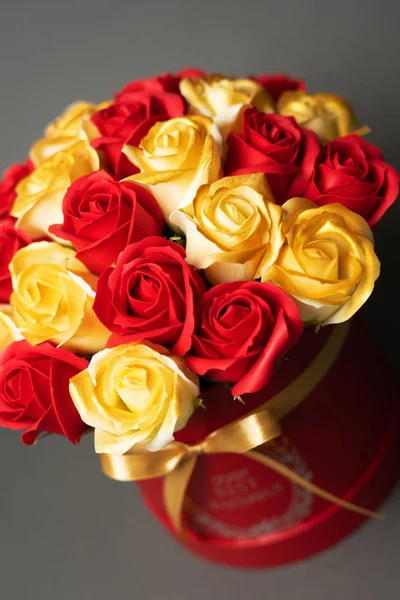 Flores en flor: Un ramo de rosas rojas y doradas en una caja redonda roja sobre un fondo gris . — Foto de Stock