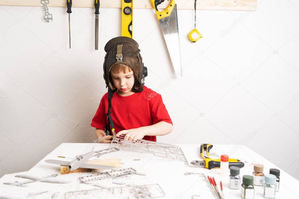Childrens hobbies: A little boy in a pilots helmet builds a large-scale model of an airplane from plastic parts using a circuit.