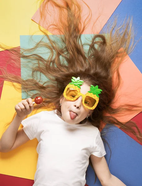 Levendige kleuren: een meisje kind met shaggy hair ligt op een veelkleurige achtergrond en likt een lolly. — Stockfoto