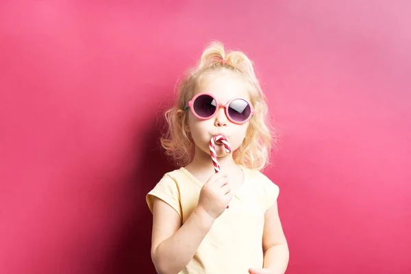 Colores brillantes: una chica con una piruleta rayada y gafas de sol sobre un fondo rosa brillante. — Foto de Stock