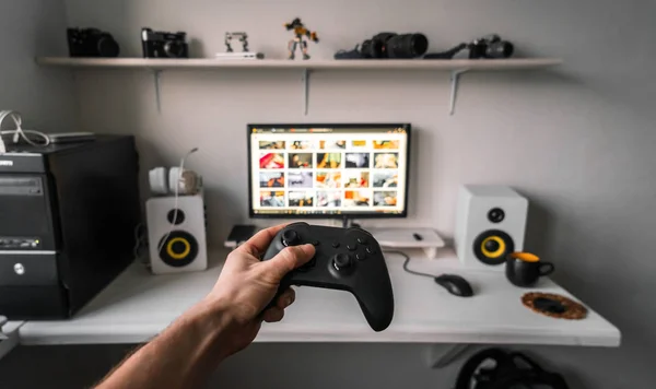 Cloud gaming: a gamepad in a male hand against the background of a gaming table and a monitor with a lot of games.