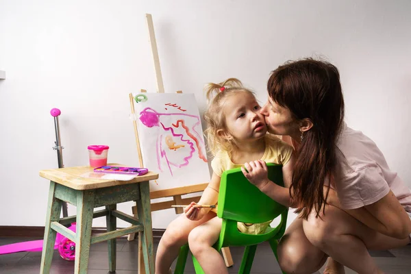Παιδική δημιουργικότητα: ένα μικρό κορίτσι ζωγραφίζει με ένα πινέλο και ακουαρέλες σε καμβά με ένα καβαλέτο. Η μαμά φιλάει το παιδί στο μάγουλο. — Φωτογραφία Αρχείου